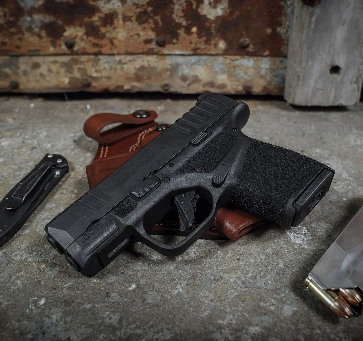 Springfield Hellcat OSP Micro-Compact pistol resting on a brown leather holster with a pocket knife placed to the left — ideal concealed carry setup.